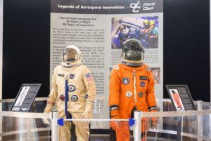 Two vintage astronaut suits are displayed behind a railing: one beige with NASA patches, the other orange. A detailed exhibit sign sits in the background.